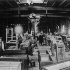 The original Matot shop, with workers working on building wooden dumbwaiters, shown in black and white.